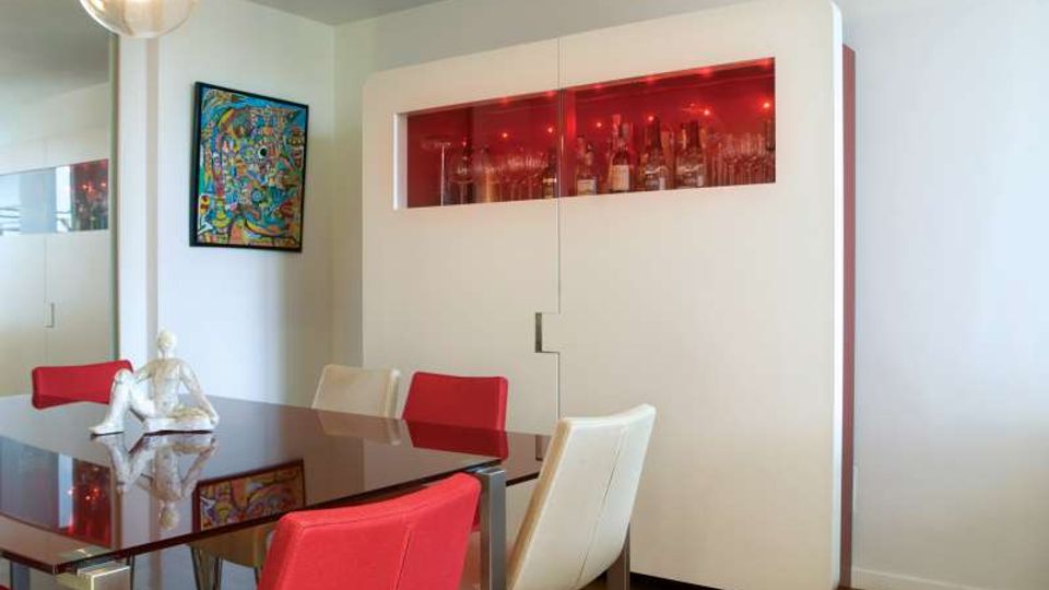 Sala da pranzo moderna con tavolo in vetro e sedie rosse e bianche, illuminata da una lampada a sospensione. Sullo sfondo un grande mobile bianco con vetrina superiore retroilluminata in rosso che espone bottiglie e bicchieri, con quadro colorato sulla parete.