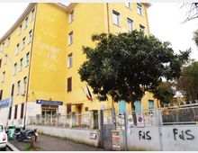Edificio scolastico a più piani con facciata gialla e finestre con persiane verdi, circondato da recinzione metallica e muro con cartelli e graffiti. Davanti all’ingresso si vedono un albero grande, un’area di accesso con cancelli e una bandiera italiana.