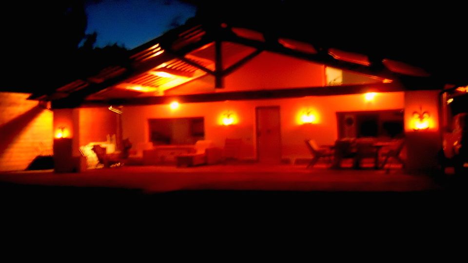Casa a un piano vista di notte con ampio portico coperto da tetto a falde e travi a vista, illuminata da luci calde sulle pareti. Davanti si vede un patio con sedie e tavolo, circondato dal buio del giardino esterno.
