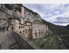 Grande complesso monastico in pietra addossato a una parete rocciosa, con lunghe facciate ad archi e un camminamento con scala e parapetto in primo piano. Sullo sfondo si vede una valle montana boscosa e cielo nuvoloso.