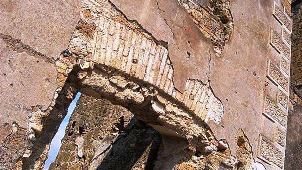 Rovine di un edificio in muratura con pareti intonacate e parti crollate, che lasciano visibili archi e aperture senza infissi. Si notano mattoni e pietre a vista, con il cielo azzurro sullo sfondo attraverso le finestre.
