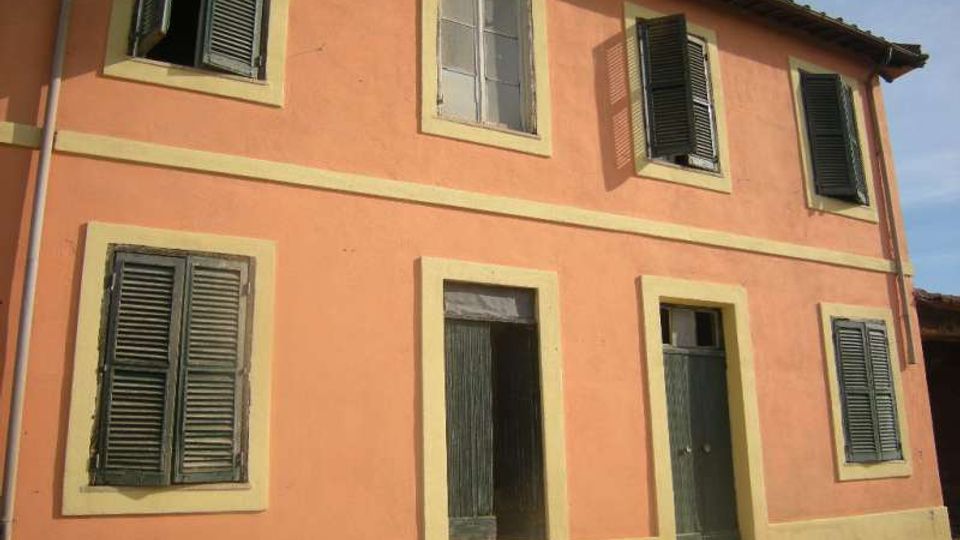Facciata di una casa a due piani intonacata color salmone con cornici beige, persiane verdi e due porte d’ingresso al piano terra con piccoli gradini. Tetto a falde con sporto e finestre disposte regolarmente lungo la parete esterna.