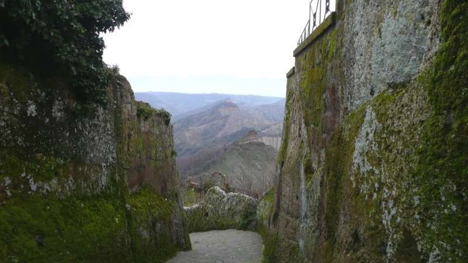 Stretto camminamento in pietra in discesa tra alte mura rocciose ricoperte di muschio e vegetazione, con una ringhiera in alto a destra. Sullo sfondo si apre una vista su colline e valli montuose.