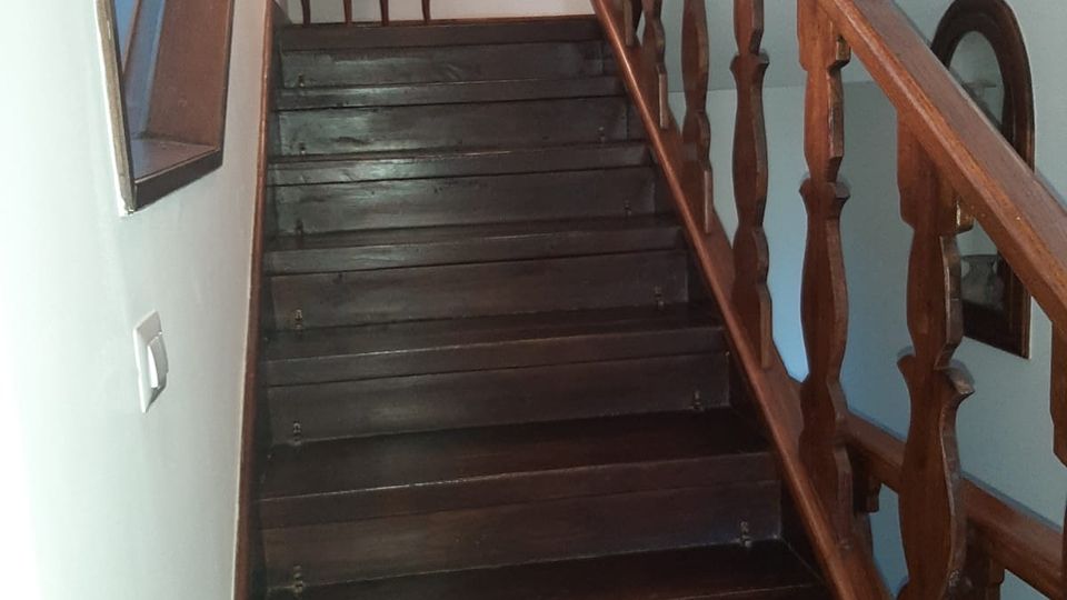 Scala interna in legno scuro con gradini lucidi e ringhiera intagliata, in un corridoio con pareti bianche. In cima alla rampa si vede un piccolo mobile a cassetti e un quadro appeso, con una finestra laterale che illumina lo spazio.