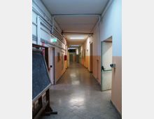 Corridoio interno lungo in un edificio scolastico o industriale, con pavimento piastrellato grigio, pareti bicolore e illuminazione a neon. Sono visibili tubazioni a vista sul soffitto, porte laterali e un cartello di uscita di emergenza illuminato.