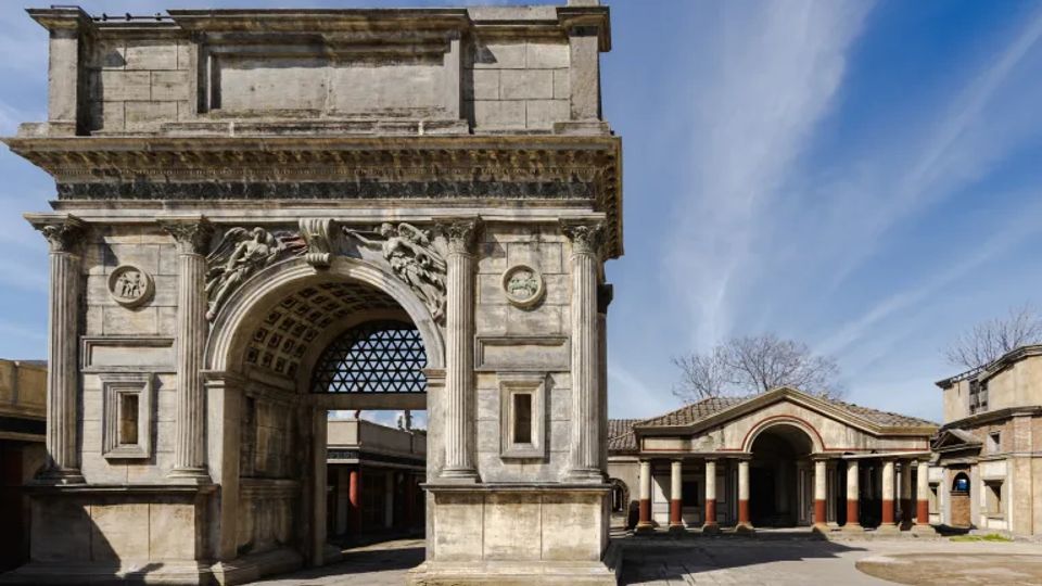 Arco monumentale in pietra con grande apertura a volta cassettonata, colonne laterali e bassorilievi decorativi sulla facciata. Davanti si apre un’ampia piazza lastricata, con sullo sfondo un portico con colonne e tetto in coppi sotto un cielo azzurro.