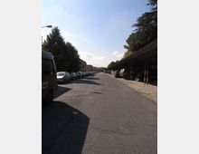 Strada asfaltata ampia in un’area industriale, con auto parcheggiate in fila sul lato sinistro e una lunga tettoia metallica con veicoli e materiali stoccati sul lato destro. Alberi e lampioni costeggiano la strada, che si estende in prospettiva verso edifici bassi sullo sfondo.