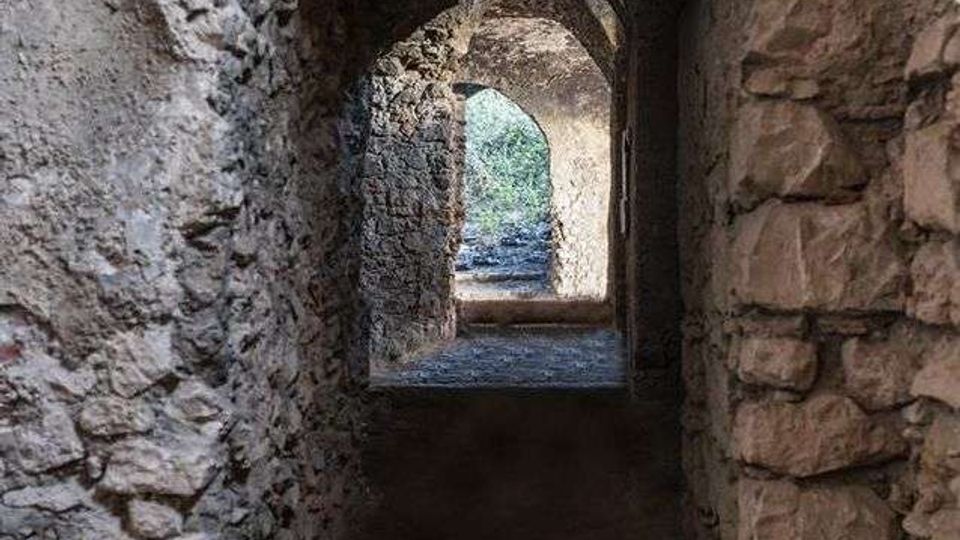 Corridoio stretto in pietra grezza con soffitto a volta e una serie di archi che conducono verso un’apertura luminosa. In fondo si intravede l’esterno con vegetazione, mentre il pavimento è in terra o pietra scura.