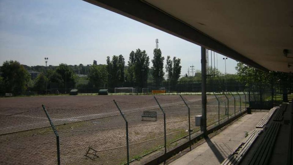Tribuna coperta di un impianto sportivo con gradinate in cemento, affacciata su un campo in terra battuta recintato con rete metallica. Sullo sfondo si vedono alberi, pali dell’illuminazione e una piccola porta da calcio.