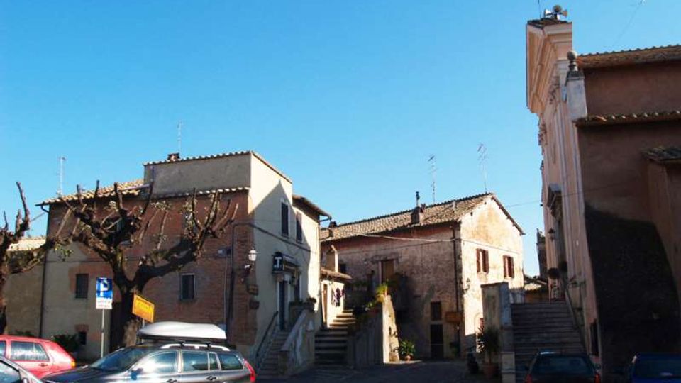 Piccola piazza in un borgo con pavimentazione in pietra e alcune auto parcheggiate, circondata da case basse in muratura con tetti in coppi. Al centro e sul lato destro si vedono scalinate che salgono tra gli edifici, con alberi potati e insegne stradali sotto un cielo sereno.