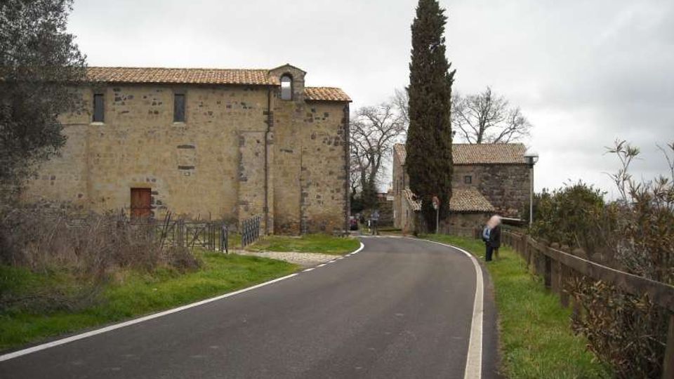 Strada asfaltata di campagna che curva tra edifici in pietra con tetti in coppi, simili a un complesso rurale o antico. Lungo la strada si vedono recinzioni, vegetazione e un alto cipresso, con alberi spogli sullo sfondo sotto cielo nuvoloso.