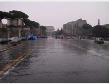 Ampio viale urbano in pavé bagnato dalla pioggia, con auto e semafori lungo la strada. Ai lati si vedono rovine e edifici storici con alberi, e in fondo un grande anfiteatro in pietra.