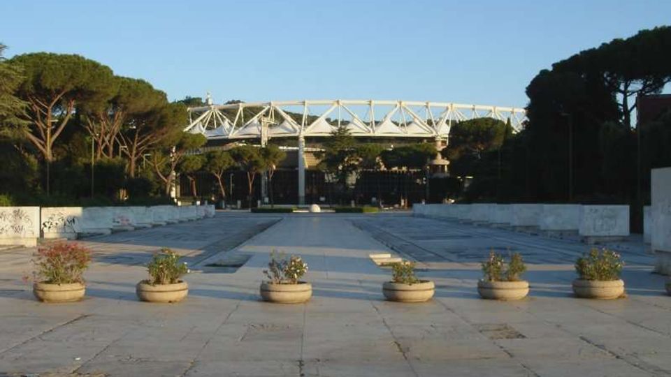 Ampia piazza pavimentata con grandi lastre di cemento e una fila di fioriere rotonde in primo piano, affiancata da muretti bassi. Sullo sfondo si vede una struttura tipo stadio con travi metalliche bianche, circondata da alti pini sotto un cielo sereno.