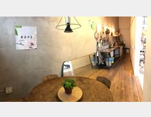 Interno di un laboratorio cucina dal design industriale, con pavimento in legno e pareti grezze. In primo piano c’è un tavolo rotondo in legno con una piccola pianta, mentre sul fondo si vede una lunga postazione con lavelli e utensili appesi, illuminata da lampade a sospensione.