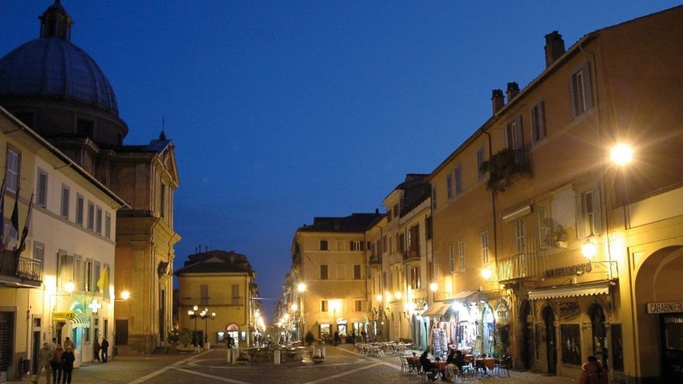 Veduta serale di una piazza urbana con edifici storici a più piani illuminati da lampioni e negozi, e tavolini all’aperto lungo la strada. A sinistra si distingue una grande chiesa con cupola che domina l’area pedonale lastricata.