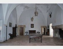 Ampia sala interna con soffitto a volte bianche e pavimento in pietra, arredata con un lungo tavolo centrale in legno e mobili scuri lungo le pareti. Sulla destra si vede un grande camino in pietra e diverse porte e archi che collegano ad altri ambienti.