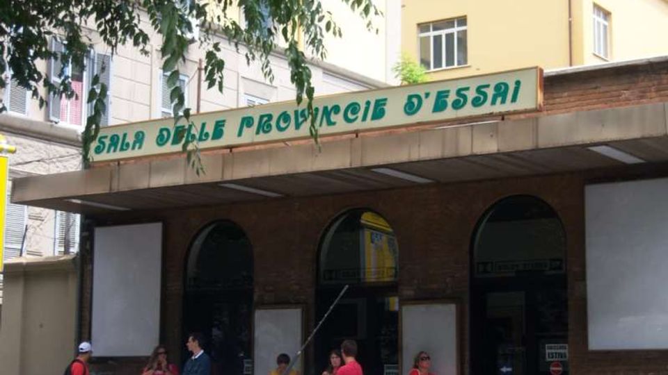 Ingresso di un cinema con insegna “Sala delle Provincie d’Essai”, tettoia sporgente e facciata in mattoni con tre grandi aperture ad arco. Sui lati dell’entrata sono presenti pannelli per locandine, con edifici residenziali sullo sfondo e rami di alberi in alto.