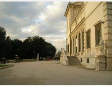 Viale pedonale accanto a un grande edificio storico in pietra chiara, con facciata monumentale, finestre con grate e una scalinata che conduce a portici ad arco. Ai lati si vedono statue su piedistalli e, sullo sfondo, un’area verde con alberi sotto un cielo nuvoloso.