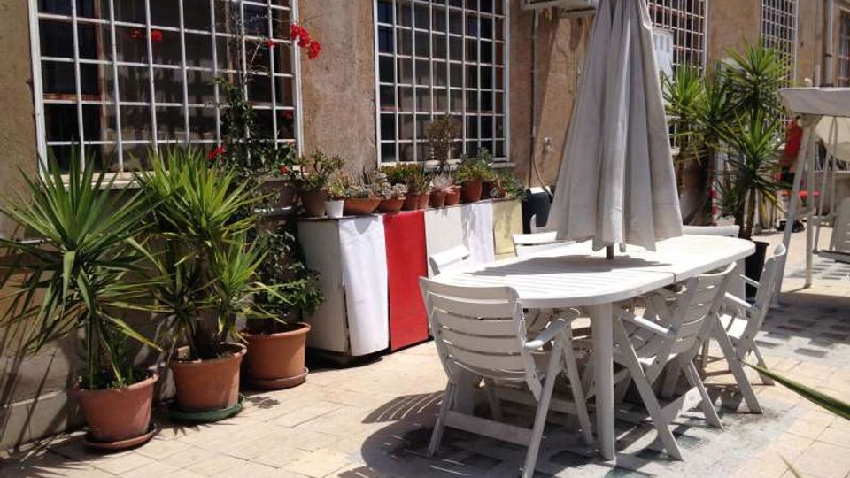 Terrazza esterna pavimentata accanto a un edificio con grandi finestre a griglia metallica, con un tavolo e sedie bianchi sotto un ombrellone chiuso. Lungo la parete ci sono numerosi vasi con piante e una fila di piccoli contenitori, oltre a un’unità di condizionamento fissata in alto.