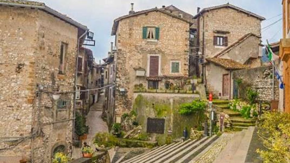 Borgo storico in pietra con case ravvicinate e facciate in muratura, finestre con scuri e tetti in coppi. In primo piano una scalinata e una strada in pendenza con gradini, piccoli muretti e aiuole, tra vicoli stretti e costruzioni addossate.