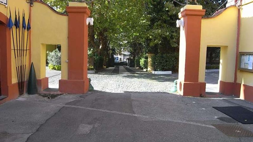 Ingresso con cancello aperto tra due pilastri intonacati giallo e rosso, sormontati da statue e collegati da un arco in ferro. Oltre il varco si vede un viale lastricato che conduce in un’area alberata.