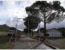 Strada asfaltata in un’area aperta con un telaio metallico bianco e rosso sopra la carreggiata, simile a un ostacolo o percorso di addestramento. Ai lati ci sono alti pini, un edificio basso con pannelli chiari e un camion parcheggiato, con colline sullo sfondo sotto un cielo nuvoloso.