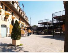Piazzale pavimentato all’aperto accanto a un edificio a più piani con balconi e arcate, con grandi fioriere e una scalinata d’ingresso. Sul lato destro si vedono strutture metalliche sopraelevate con ringhiere, simili a tribune o passerelle, sotto un cielo sereno.