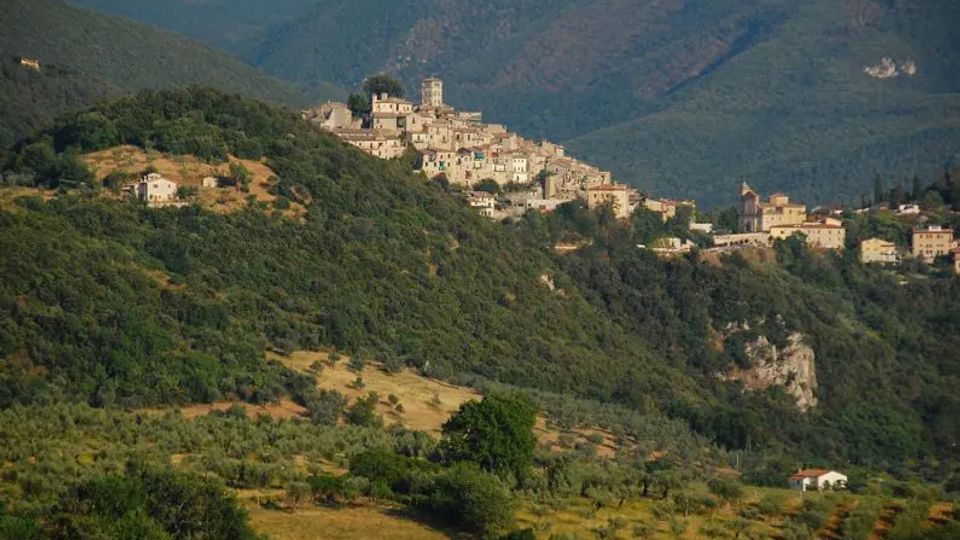 Borgo medievale arroccato su una collina, con case in pietra e una torre campanaria, circondato da pendii boscosi e terrazzamenti agricoli. Sullo sfondo si vedono montagne verdi e una valle ampia.