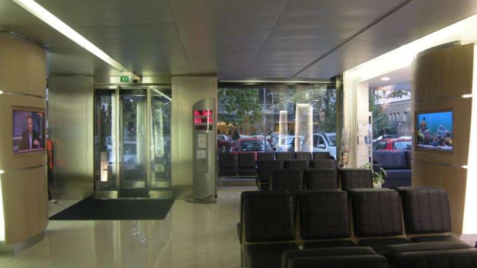 Interno di una hall d’ingresso con pavimento lucido, file di sedute nere e pareti con schermi informativi; sul fondo si vede una porta girevole in vetro e grandi vetrate affacciate sulla strada con auto parcheggiate.