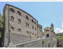 Complesso storico in pietra su un pendio, con grande edificio a più piani e finestre ad arco, collegato da una rampa con parapetto metallico. A destra si vede una chiesa in pietra con campanile e facciata semplice, sullo sfondo colline verdi e cielo sereno.