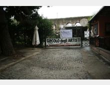 Ingresso con cancello in ferro e strada in ciottoli, con un grande striscione “Circolo degli Artisti” appeso davanti. Ai lati ci sono alberi e un ombrellone chiuso, e sullo sfondo si intravede un muro in pietra con arcate.