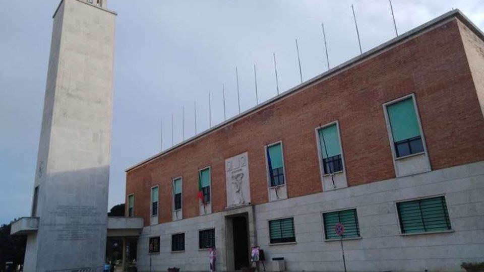 Edificio civico a due piani con facciata in mattoni rossi e basamento in pietra chiara, con ingresso centrale e finestre rettangolari. A sinistra si innalza una torre campanaria moderna in cemento chiaro, in una piazza aperta sotto un cielo nuvoloso.