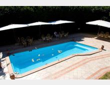 Piscina rettangolare all’aperto con bordo in pietra e pavimentazione in piastrelle geometriche, circondata da un’area solarium. Lungo un lato ci sono grandi ombrelloni bianchi con tavolini e sedie, con una siepe alta sullo sfondo.