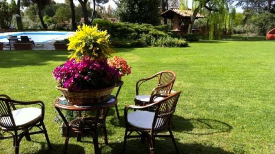 Giardino con ampio prato verde e area salotto con tavolino e sedie in vimini, con un cesto di fiori colorati al centro. Sullo sfondo si vedono una piscina, alberi alti e una piccola struttura con tetto in tegole tra la vegetazione.