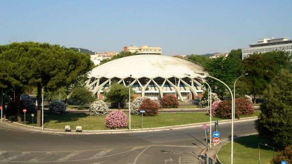 Edificio moderno a cupola bianca con struttura reticolare visibile alla base, circondato da alberi e aiuole fiorite. In primo piano si vede una grande rotatoria stradale con corsie e segnaletica, sullo sfondo altri edifici urbani.