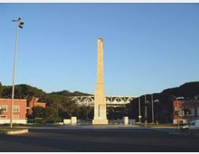 Rotonda stradale con un alto obelisco in pietra al centro, circondata da carreggiate e pali dell’illuminazione. Sullo sfondo si vedono edifici bassi, alberi e una struttura metallica simile a un ponte sotto un cielo sereno.