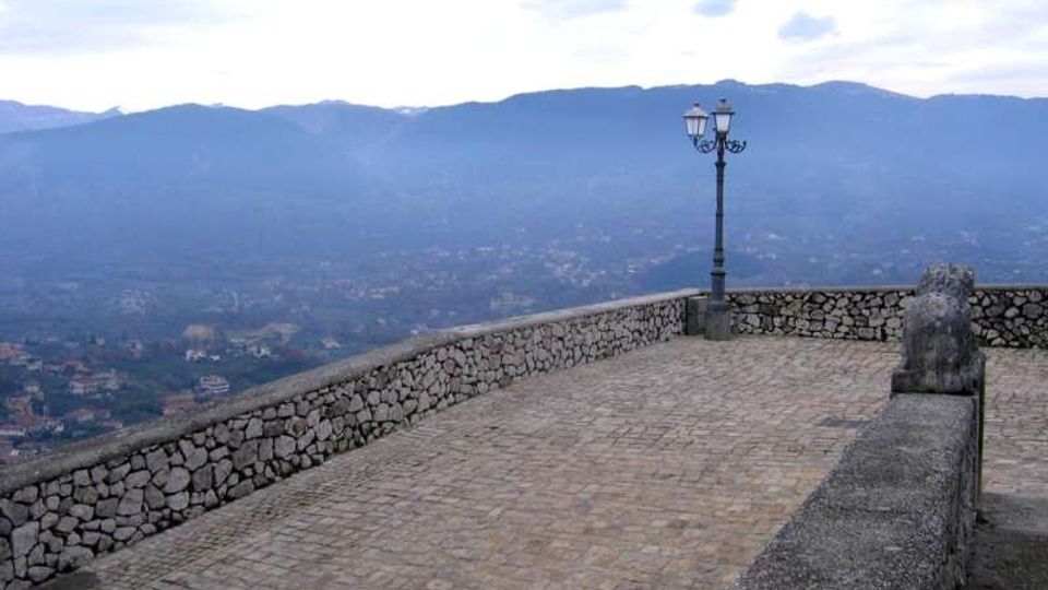 Terrazza panoramica in pietra con pavimentazione lastricata e parapetti in muratura, con un lampione a due luci vicino al bordo. Sullo sfondo si vedono una valle con abitazioni sparse e una catena montuosa sotto un cielo nuvoloso.