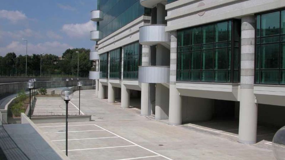 Parcheggio esterno con stalli segnati a terra accanto a un moderno edificio per uffici con facciata in vetro e pilastri in cemento. Sul lato del fabbricato si vedono balconate semicircolari e lampioni lungo il percorso, con alberi sullo sfondo.