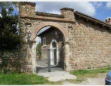 Ingresso in pietra di un edificio rurale con grande arco in mattoni e cancello in ferro battuto, inserito in un muro di pietra con merlature e lampioni ai lati. Sullo sfondo si vede il cortile con un percorso in cemento e una facciata in pietra sotto un cielo parzialmente nuvoloso.