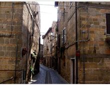 Stretta via acciottolata tra alti edifici in pietra, con persiane alle finestre, vasi di fiori alle pareti e lampioni lungo il percorso. Cavi e tubazioni corrono sulle facciate, mentre la strada curva e si restringe verso il fondo.