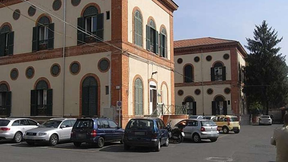 Edificio a più piani in stile storico-industriale con facciata chiara, cornici in mattoni rossi e numerose finestre ad arco e oculi circolari. Davanti si trova un ampio parcheggio asfaltato con auto, con un secondo corpo di fabbrica sullo sfondo e un albero alto sul lato destro.