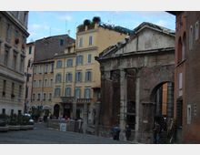 Piazza urbana con pavimentazione in pietra, circondata da palazzi storici dai toni giallo e beige con finestre regolari. In primo piano si vedono i resti di un’antica struttura in mattoni con colonne e frontone parzialmente crollato, delimitata da transenne.