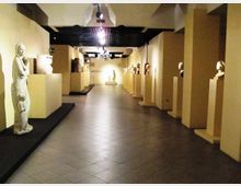 Corridoio interno di un museo o galleria con pavimento in piastrelle e illuminazione a soffitto, fiancheggiato da piedistalli che espongono sculture e busti. Le pareti chiare e la prospettiva lunga conducono verso un’area espositiva sul fondo.