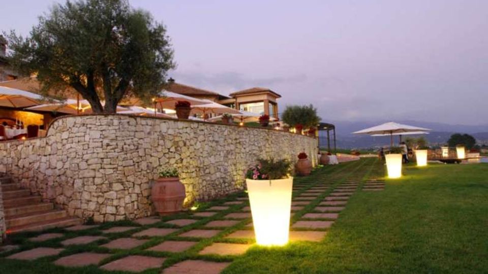 Terrazza all’aperto con muretto in pietra e grande ulivo, affacciata su un prato con percorso di lastre quadrate. Lungo il vialetto sono presenti vasi luminosi e ombrelloni bianchi, con vista su colline sullo sfondo al crepuscolo.