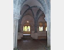 Interno di un chiostro in pietra con colonne e archi ogivali che sorreggono volte a costoloni. Sul fondo, una breve scalinata conduce a una loggia con arcate aperte verso un cortile verde.