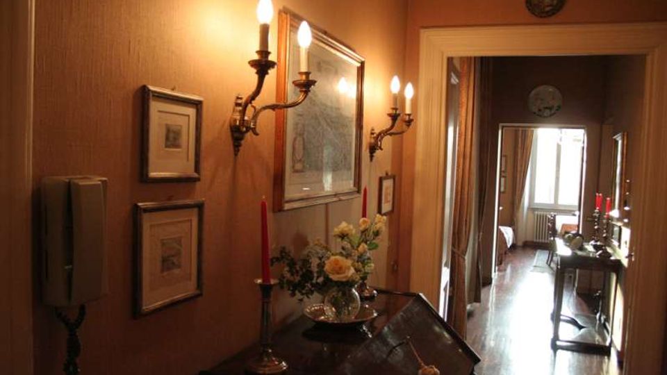 Corridoio interno con pareti color ocra illuminate da applique a candelabro, quadri incorniciati e un mobile scuro con vassoio, fiori e candelabri. In fondo si apre una stanza con pavimento in legno, finestra luminosa e un tavolino d’appoggio vicino all’ingresso.