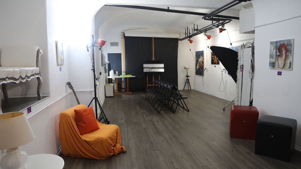 Sala espositiva con pareti bianche e pavimento in legno, con quadri appesi e file di sedie nere rivolte verso un fondale nero con cavalletto. Sono presenti luci da studio e un grande ombrello fotografico, oltre a una poltrona con coperta arancione e piccoli tavolini.