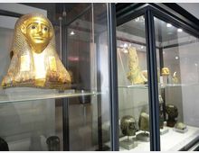 Vetrine museali in vetro e metallo che espongono reperti egizi, tra cui una maschera funeraria dorata e varie teste scultoree su piedistalli. L’ambiente interno è illuminato da faretti e presenta scaffali trasparenti con oggetti disposti su più livelli.