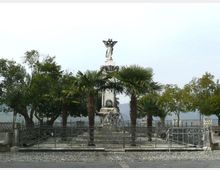 Monumento in pietra con statua sulla sommità, al centro di una piccola area recintata con cancellata in ferro battuto e pavimentazione in ciottoli. Intorno ci sono palme e altri alberi, con vista sul lago e una balaustra sullo sfondo.