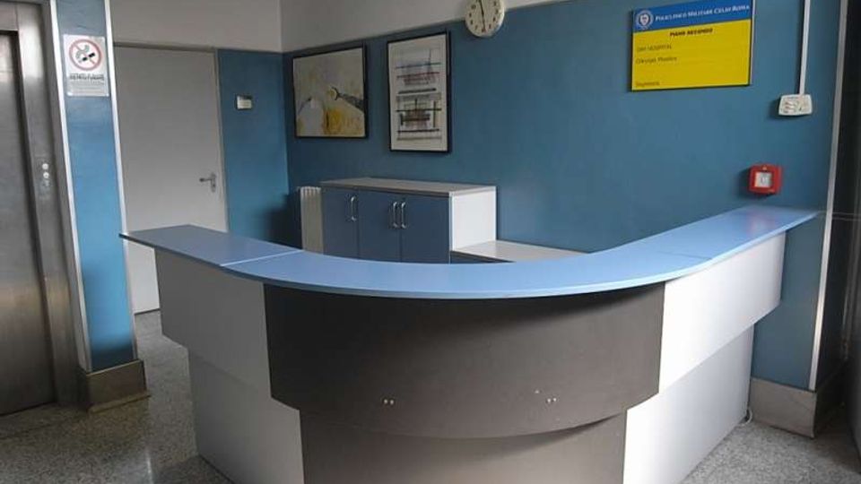Reception di un ufficio con bancone curvo bianco e blu e pareti azzurre. Sullo sfondo si vedono una porta, un armadietto, quadri e una bacheca con orologio e dispositivi di sicurezza, con pavimento in graniglia.